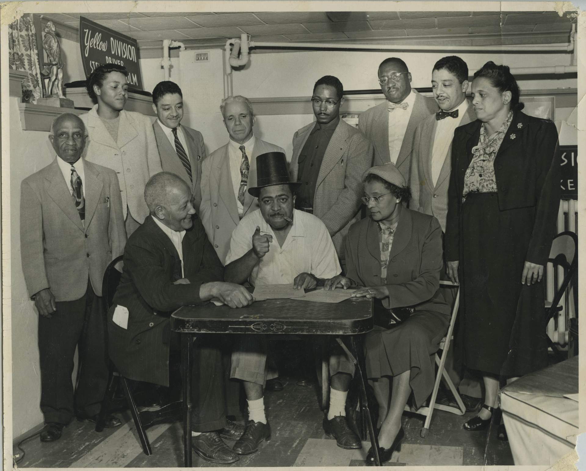 Brent, man in top hat [Count Basie?], and woman at table, 8 people standing behind them